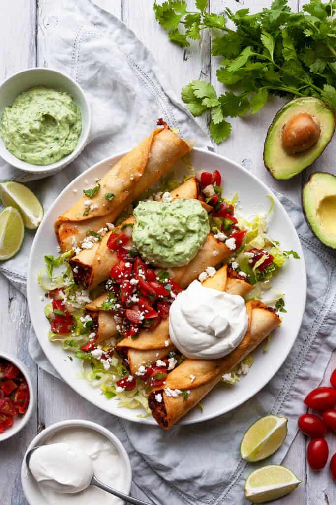 plate of chicken flautas on a bed of lettuce topped with avocado crema, sour cream, and pico de gallo