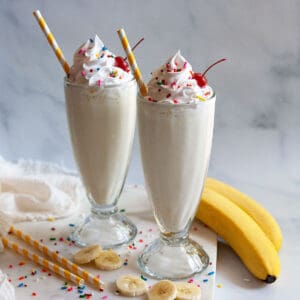 two banana milkshakes in old fashioned milkshake glasses topped with sprinkles and cherries