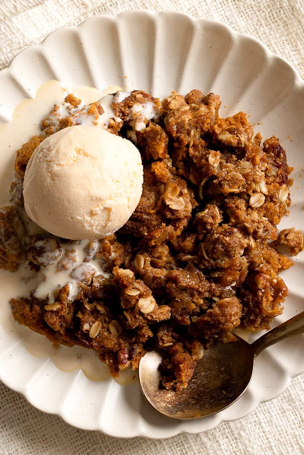 Close-up view of warm pumpkin crisp topped with a scoop of vanilla ice cream, showing the crunchy oat topping and creamy melted ice cream on a scalloped plate.