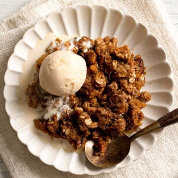 A plate of warm pumpkin crisp topped with a scoop of vanilla ice cream, set on a textured cream cloth with a spoon resting on the side.