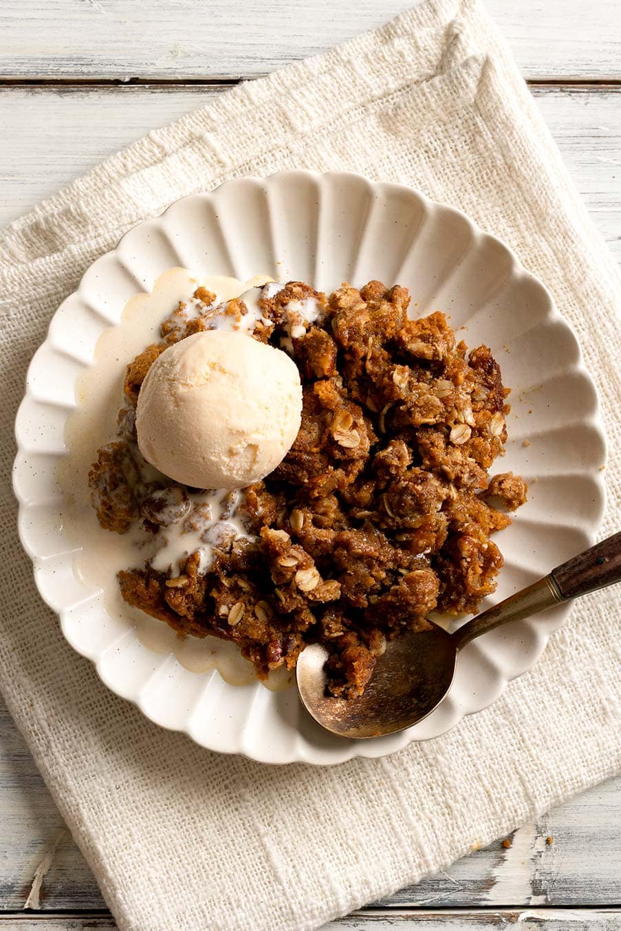 A plate of warm pumpkin crisp topped with a scoop of vanilla ice cream, set on a textured cream cloth with a spoon resting on the side.
